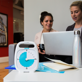 The Time Timer PLUS 20 Minute visual timer is sitting on a table in an office setting. The bright blue disk is set at a 15 minute duration. Next to the timer, on the table is a water bottle and a notepad. In the background are two women sitting on a couch, talking and looking at a laptop. 
