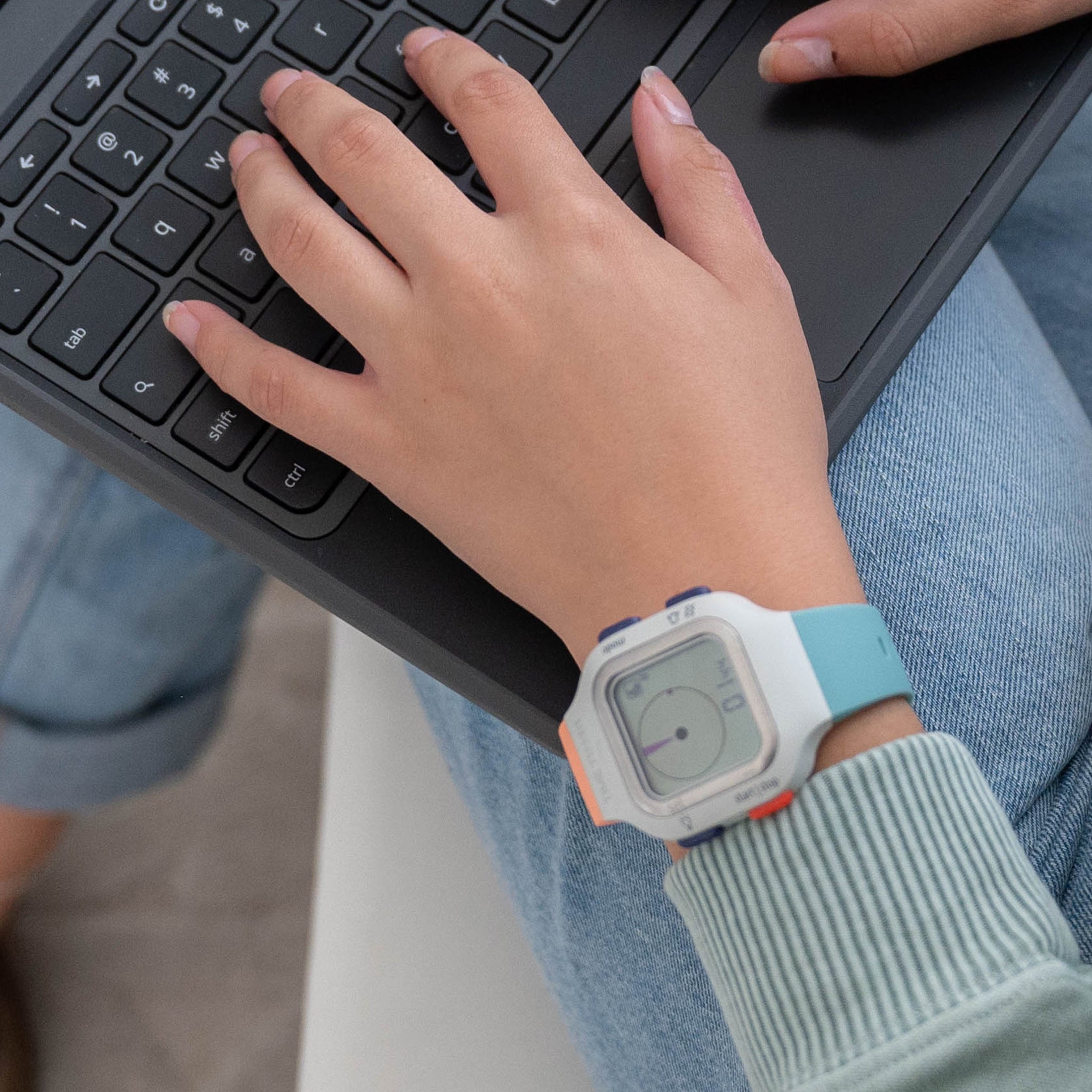 A young woman's hand is shown typing on a laptop. On her wrist is the Time Timer Watch. The watch face is the standard Arctic White option, and she has paired it with two different band colors - on top is the Sedona Orange band, on the bottom is the Caribbean blue band.  