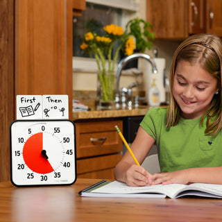 Time Timer Originals Accessory - Extra Dry Erase Activity Cards are sitting on top of the Time Timer Original 8" timer. The set is sitting next to a young girl doing her homework. The Dry Erase Activity Cards show a first/then schedule. The first card reads "First" with a drawing of a book and pencil. The second card reads "Then" with a drawing of stick figures playing with a ball.  